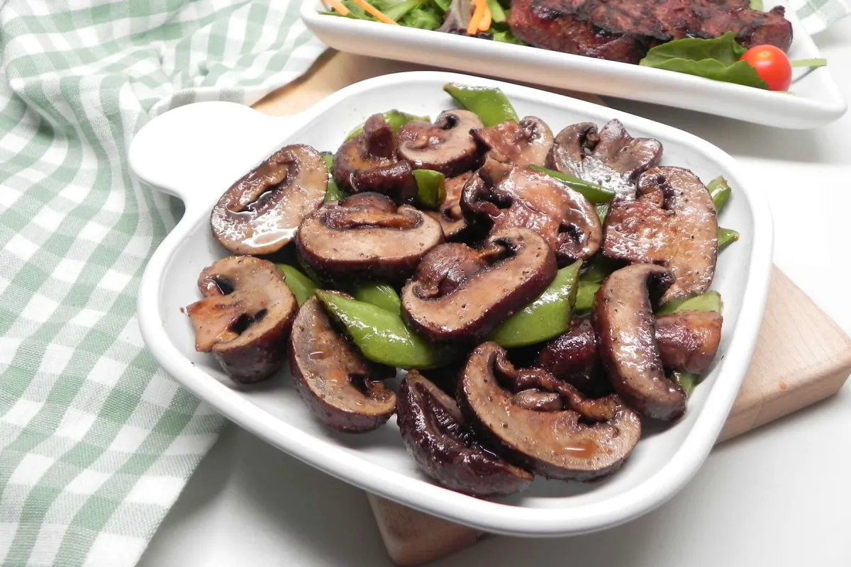 A plate of air-fried mushrooms and snap peas, with the mushrooms appearing browned and glossy, possibly seasoned. In the background, another dish contains grilled meat or salad with greens and cherry tomatoes.