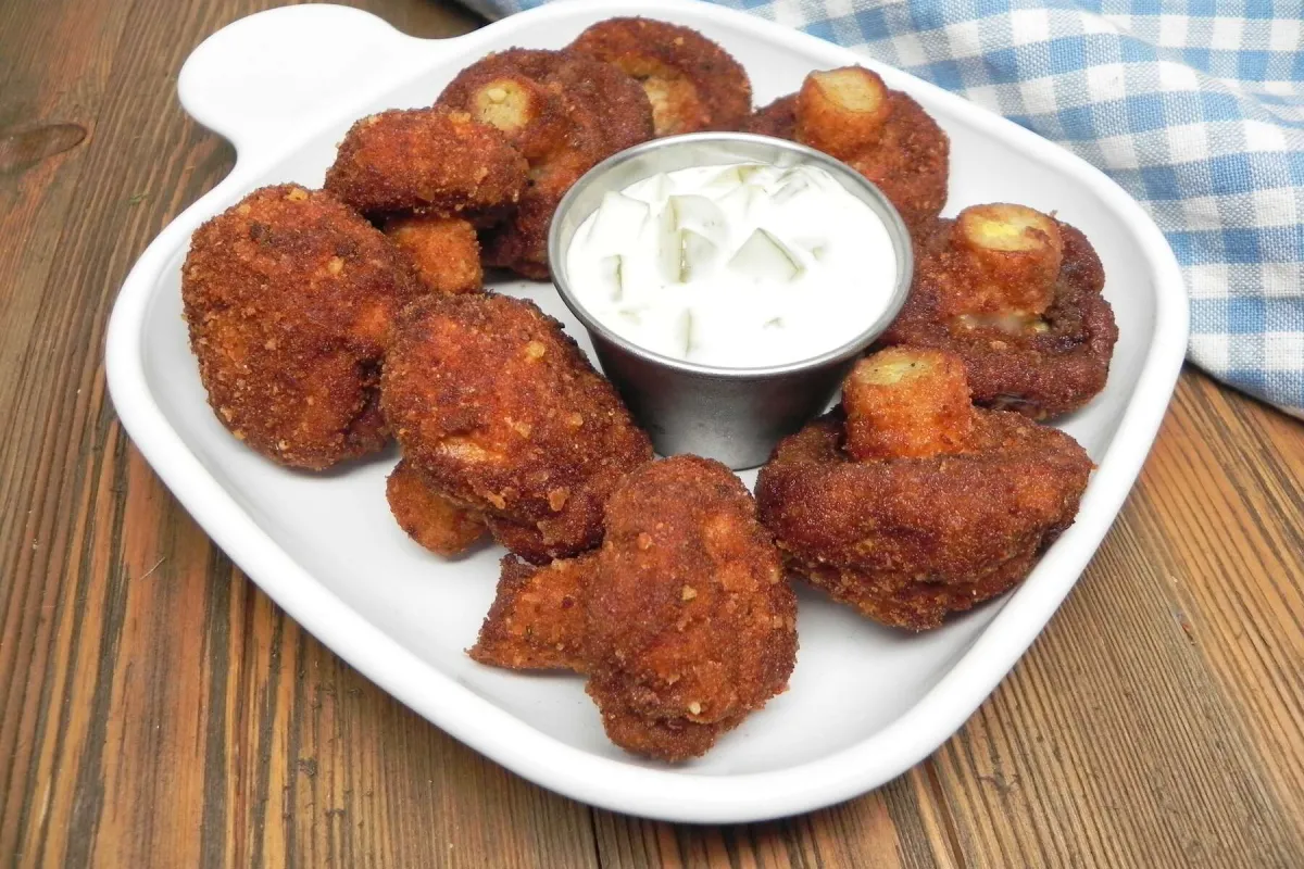 The image shows a plate of crispy, deep-fried mushrooms, golden brown in color, served with a small cup of dipping sauce, likely ranch or a similar creamy dip with chopped herbs.